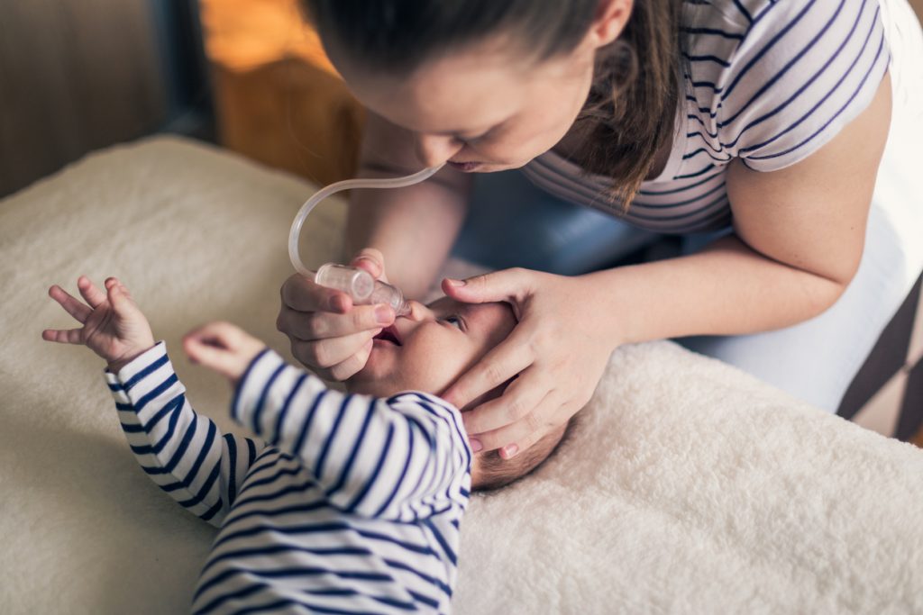 Young single mother cleans a nose of a two months old baby boy on the bed using the nose aspirator. They are dressed in matching white and blue striped clothes.