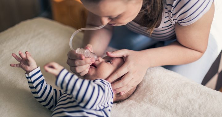 Young single mother cleans a nose of a two months old baby boy on the bed using the nose aspirator. They are dressed in matching white and blue striped clothes.